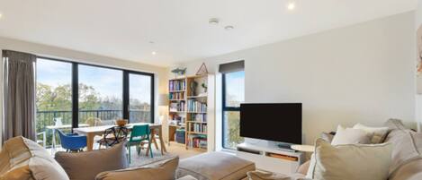 A modern living room with a large flat-screen TV, a bookshelf, and a balcony with outdoor seating.