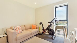 A modern living room with a sofa, a standing exercise bike, and a large window. - Lovely Luminous 3 Bed Flat, With Secure Ev Parkng (London)