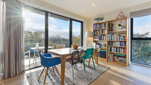 A modern living room with a large bookshelf, a dining area with a table and chairs, and a view through large windows. - Lovely Luminous 3 Bed Flat, With Secure Ev Parkng (London)