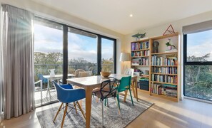 A modern living room with a large bookshelf, a dining area with a table and chairs, and a view through large windows. - Lovely Luminous 3 Bed Flat, With Secure Ev Parkng (London)
