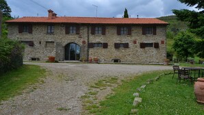 Un bâtiment en pierre avec un toit recouvert de tuiles rouges, une allée en gravier et des espaces de repos aménagés à l’extérieur.