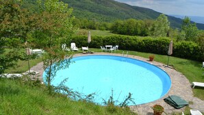 Une piscine circulaire bordée de pierres, entourée de chaises longues blanches et de parasols, le tout se fondant dans un décor de verdure luxuriante et de collines.