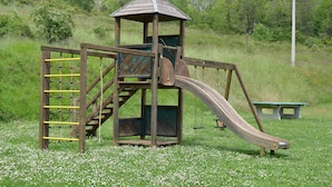 Een houten speelconstructie met een glijbaan en klimgedeelte, omgeven door gras en bloemen.