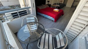 Ein Balkon mit Glastisch und Stühlen, ein Bett mit roter Bettdecke, ein Hängesessel und eine Küchenzeile.
