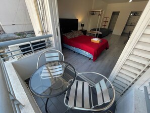 A balcony with a glass table and chairs, a bed with a red bedspread, a hanging chair, and a kitchen area.