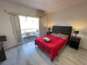 A bedroom with a bed, a nightstand, a lamp, a balcony with a table and chairs, and an air conditioning unit.
