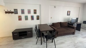 Ein modernes Wohnzimmer mit einem braunen Sofa, einem Esstisch mit Stühlen, einem Fernseher auf einem Schrank und verschiedenen Wanddekorationen.