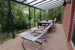 Een overdekt terras met een tafel en stoelen, omringd door weelderige begroeiing.