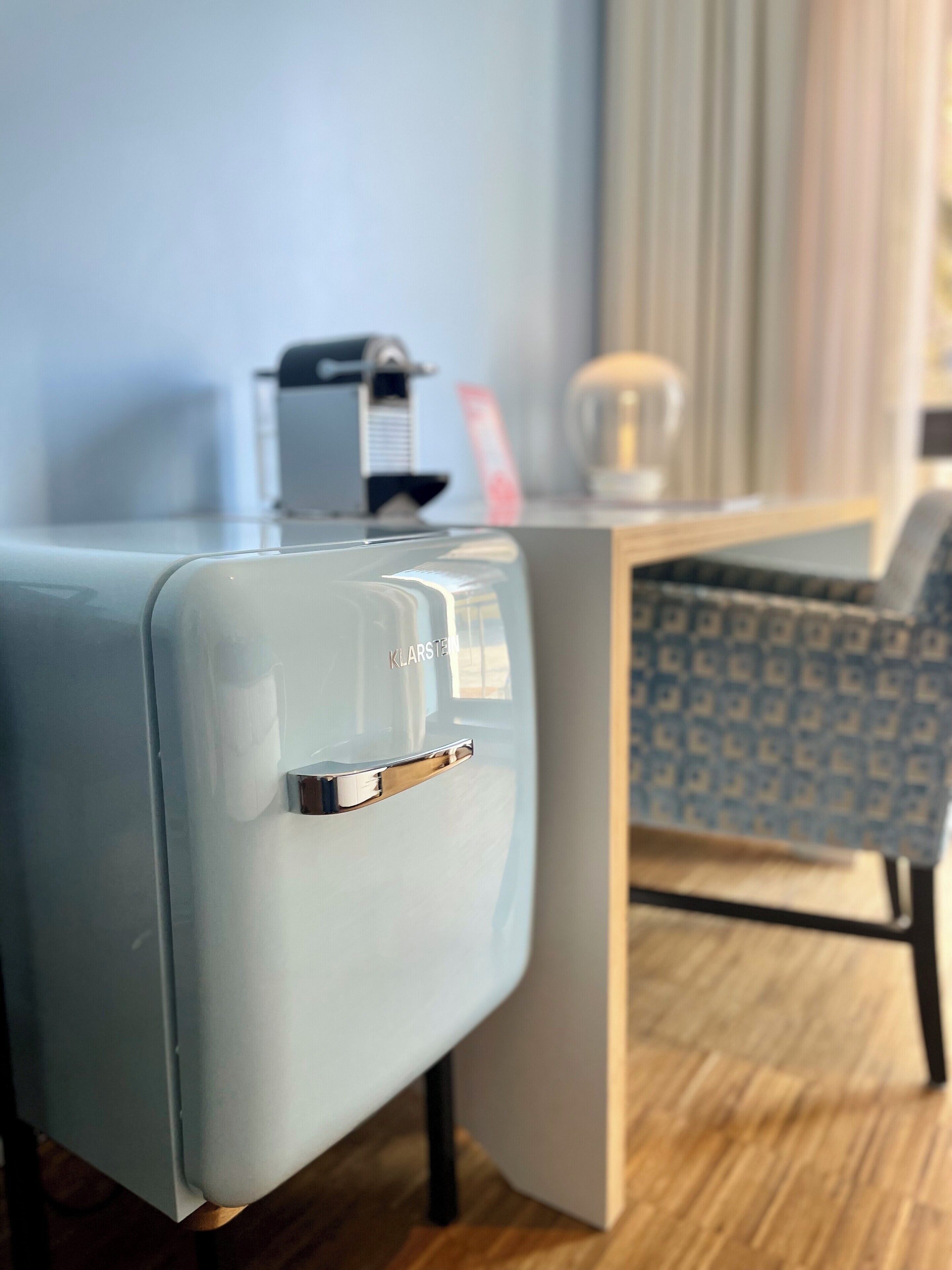A modern hotel room with a light blue suitcase, a small table with a coffee maker, and a patterned chair.