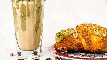 A glass of iced coffee with a creamy top and a croissant with a drizzle of white icing on a plate with scattered pistachios.