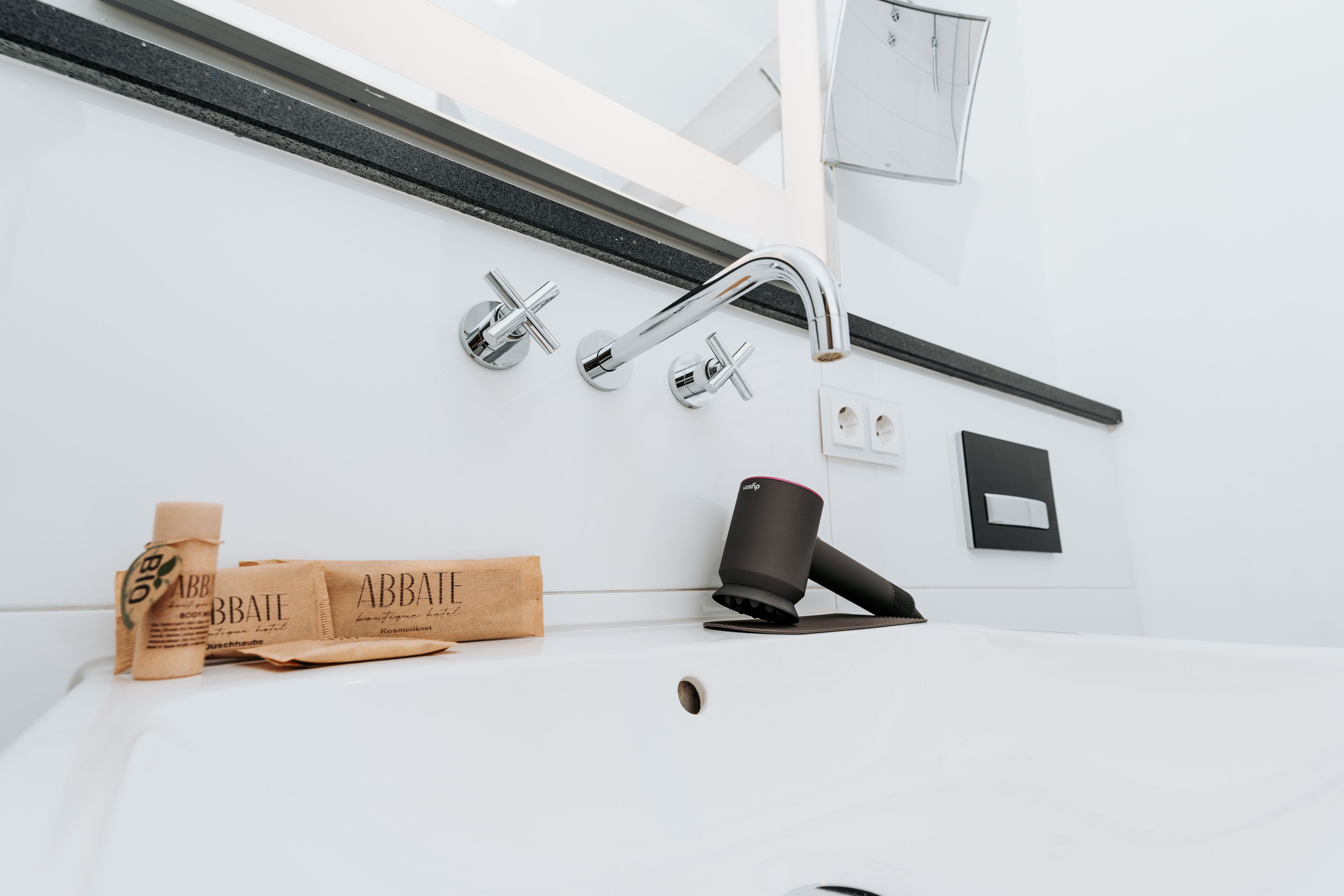 A bathroom with a white sink, two chrome faucets, a black towel holder, and a paper towel dispenser.
