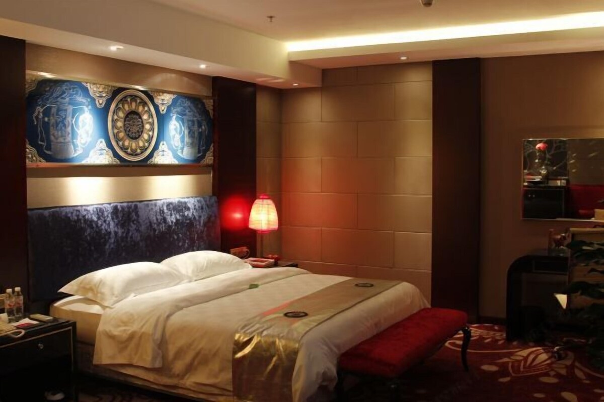 A hotel room with a large bed, a red bench, a nightstand with a lamp, and a wall with a decorative panel.