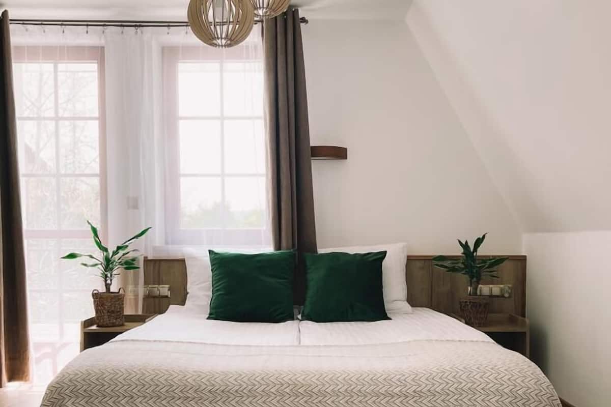 Ein Schlafzimmer mit einem großen Bett, Holzboden, zwei Nachttischen mit Blumentöpfen, einem Fenster mit Vorhängen und einer schrägen Decke mit Pendelleuchten.