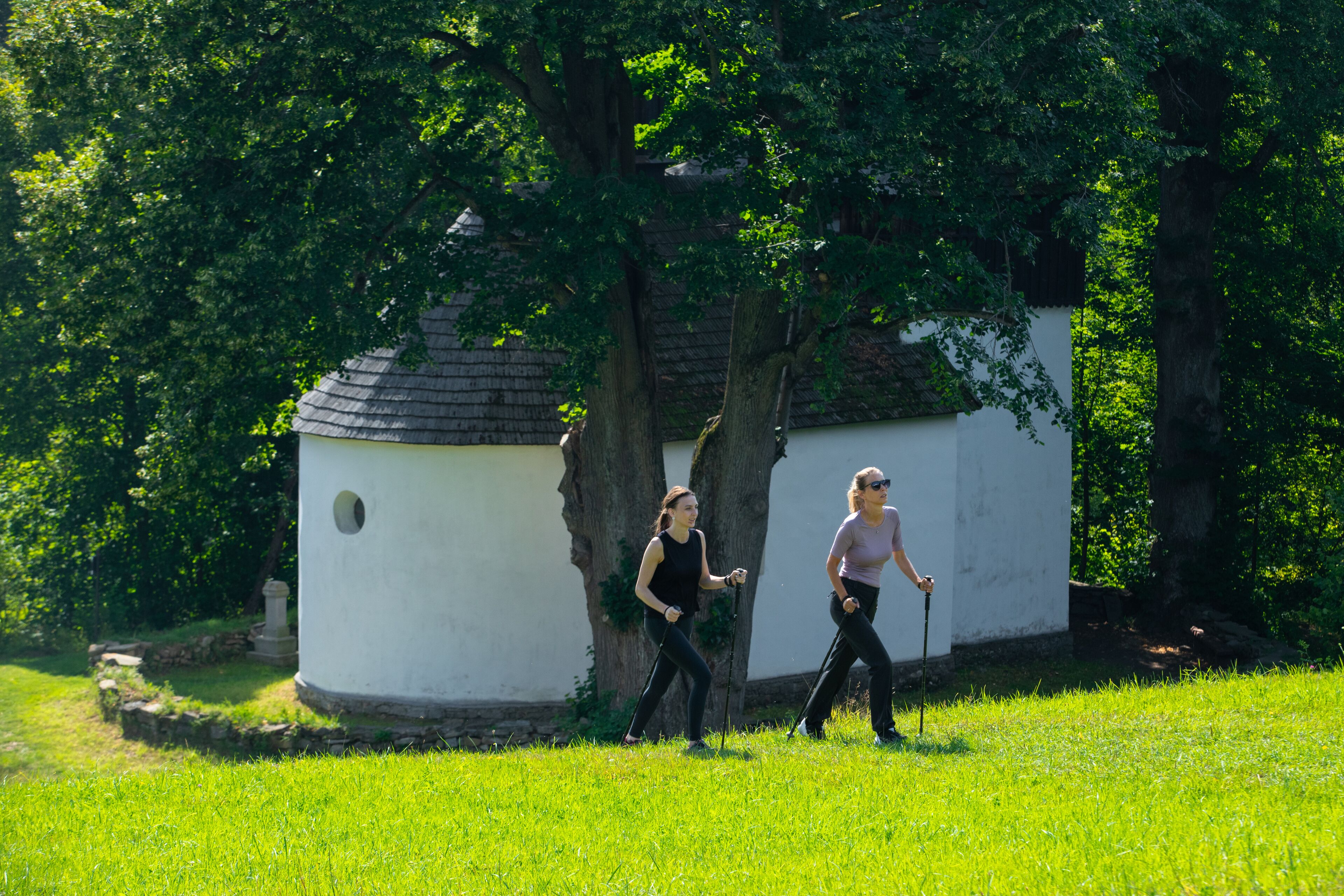 Dos personas caminando por un área con césped, árboles y un pequeño edificio blanco con una ventana circular.