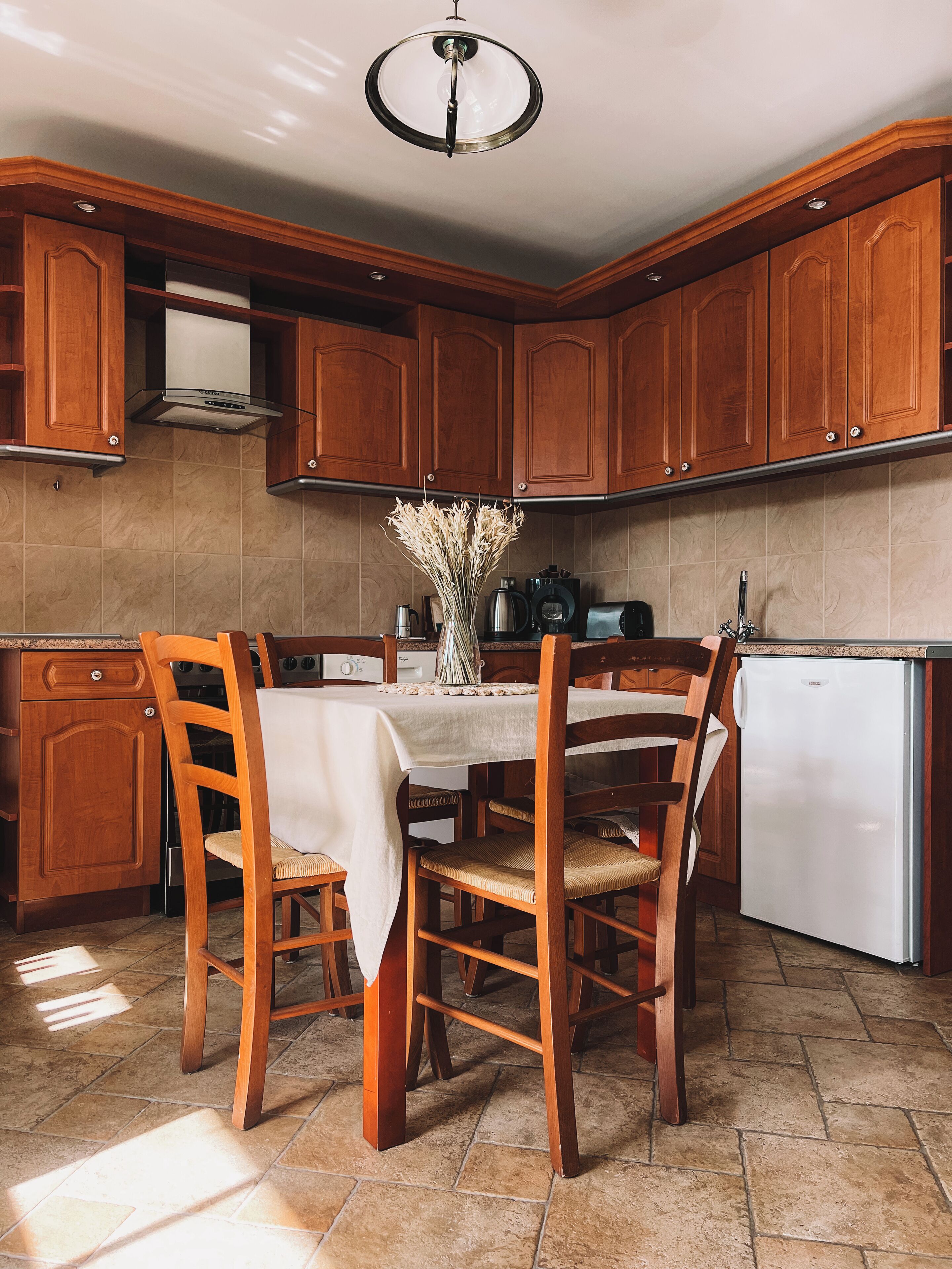A kitchen with wooden cabinets, a dining table with chairs, a refrigerator, and a ceiling light fixture.