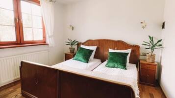 Ein Schlafzimmer mit einem hölzernen Bett, weißer Bettwäsche, grünen Kissen, einem Fenster mit Vorhängen, einem Heizkörper und einer Kristallleuchter.