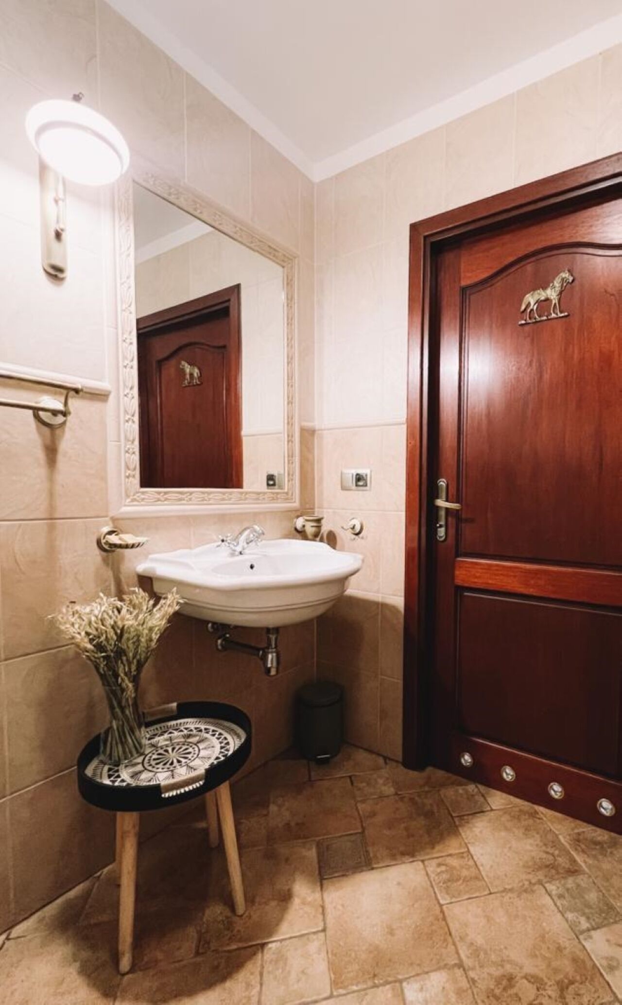 Un baño con un lavamanos blanco, una puerta de madera con un emblema de lobo, un espejo sobre el lavamanos y un taburete pequeño con un adorno.