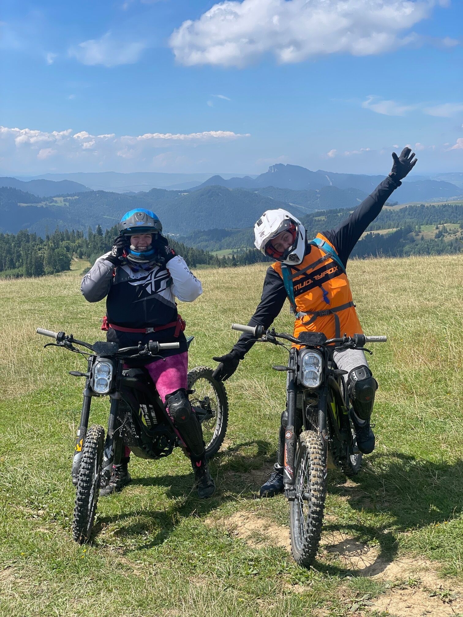 Two people on motorcycles in a grassy field with a mountainous backdrop.