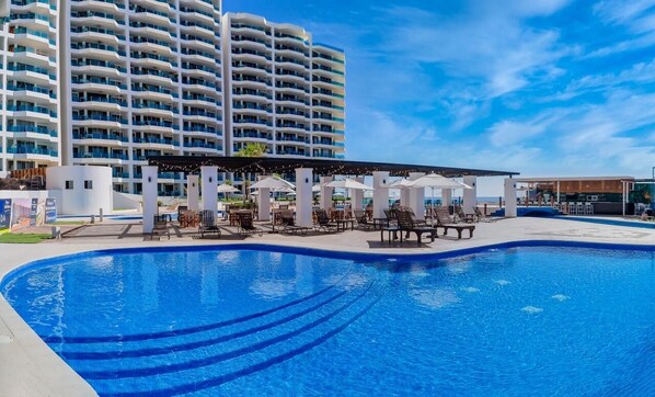 A swimming pool with lounge chairs and umbrellas, a covered seating area, and a high-rise building in the background.