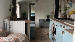 A compact kitchen with a stove, sink, and cabinets. A bed with a patterned blanket is in the foreground. - Converted French bus With Stunning Views (Beaworthy)