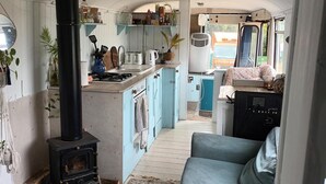 A compact kitchen with a wood-burning stove, a sink, and a stove top oven. There is a blue sofa in the foreground and a window with a view of the outside. - Converted French bus With Stunning Views (Beaworthy)