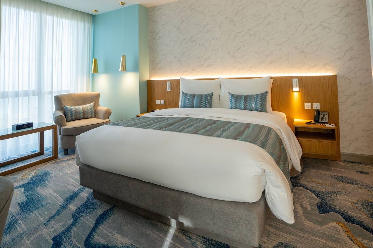 A hotel room with a large bed, a chair, a bedside table with a phone, and a window with curtains.