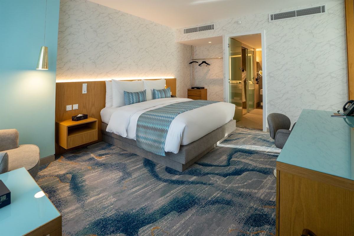 A hotel room with a bed, bedside tables, a desk, a chair, a mirror, and a bathroom visible through an open door.