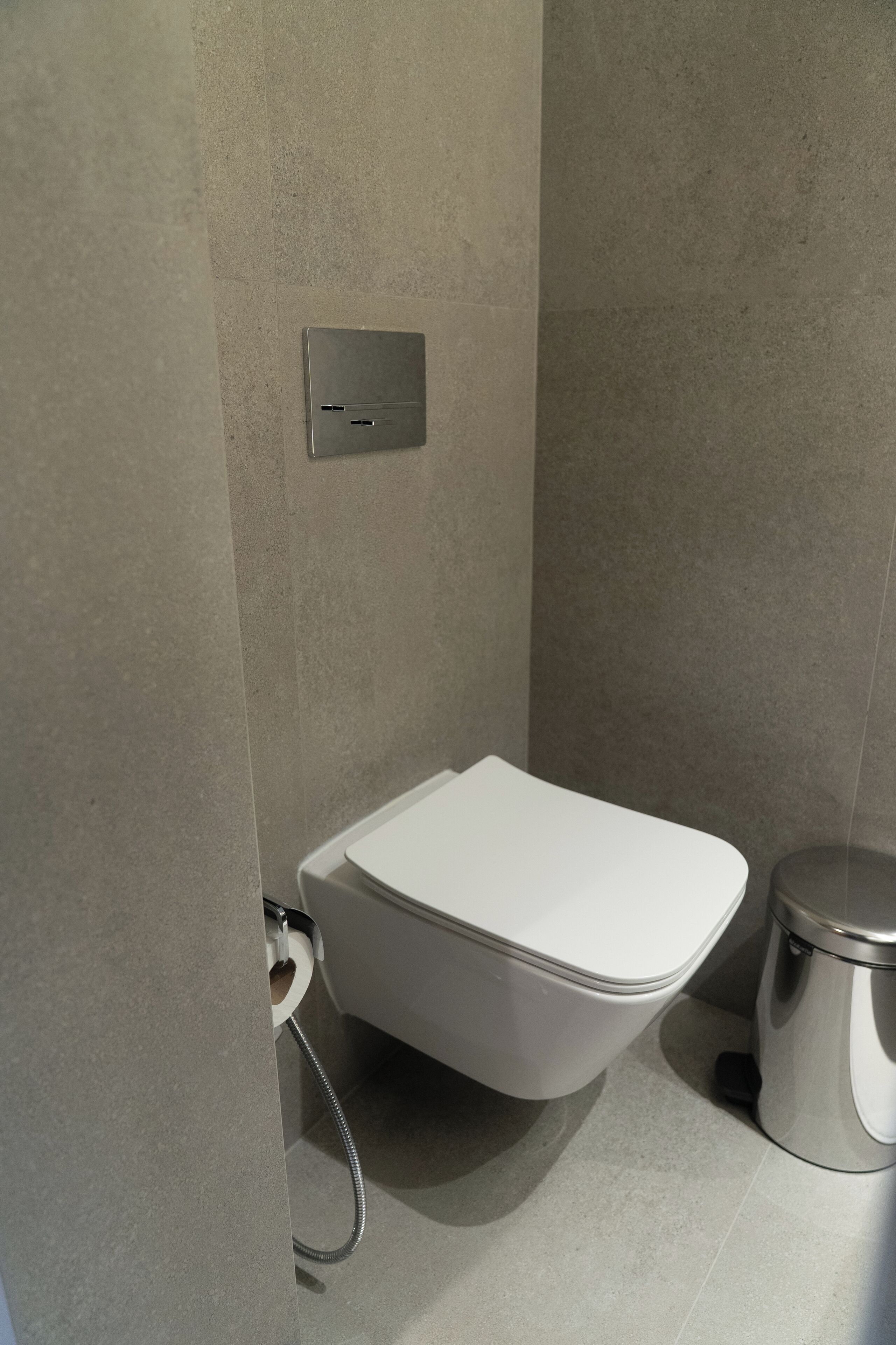 A modern bathroom with a white toilet, a wall-mounted flush handle, and a trash bin.