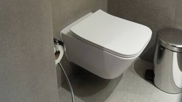 A modern bathroom with a white toilet, a wall-mounted flush handle, and a trash bin.