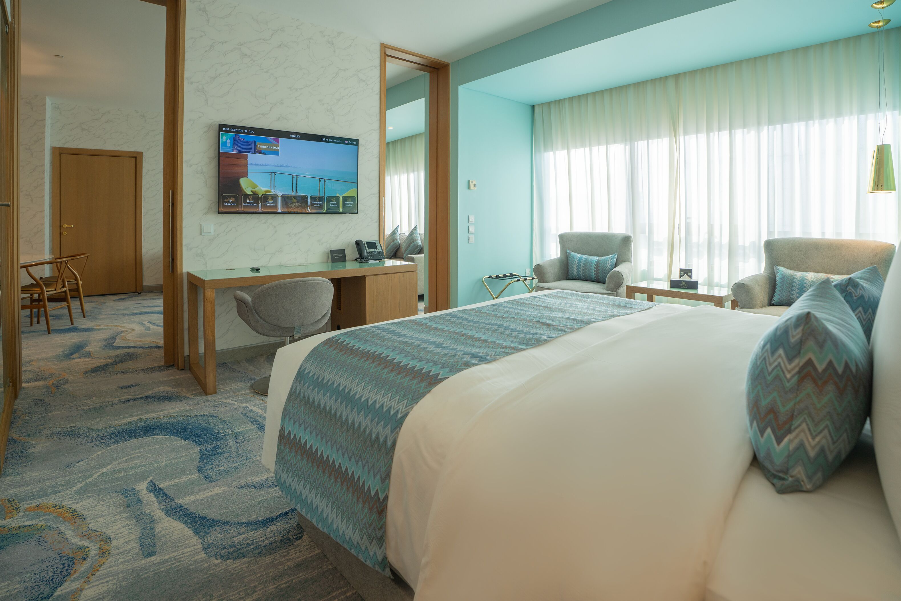 Habitación de hotel con cama, escritorio, silla, televisor y un amplio ventanal con cortinas.