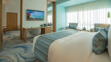 Habitación de hotel con cama, escritorio, silla, televisor y un amplio ventanal con cortinas.