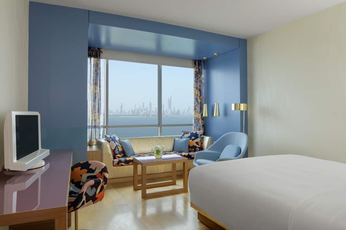 A hotel room with a large bed, a desk with a computer, a sofa with colorful cushions, a chair, and a view of a city skyline through large windows.