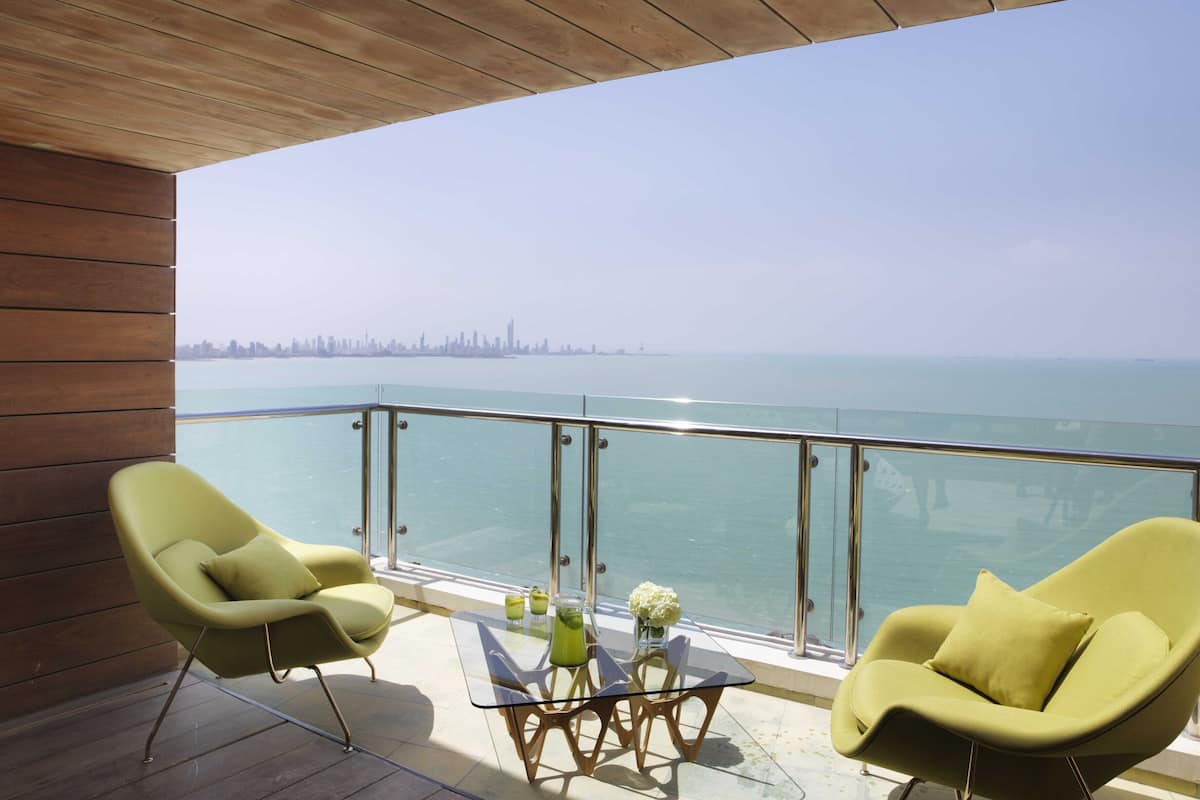 A balcony with two armchairs and a glass table, overlooking a city skyline and the sea.