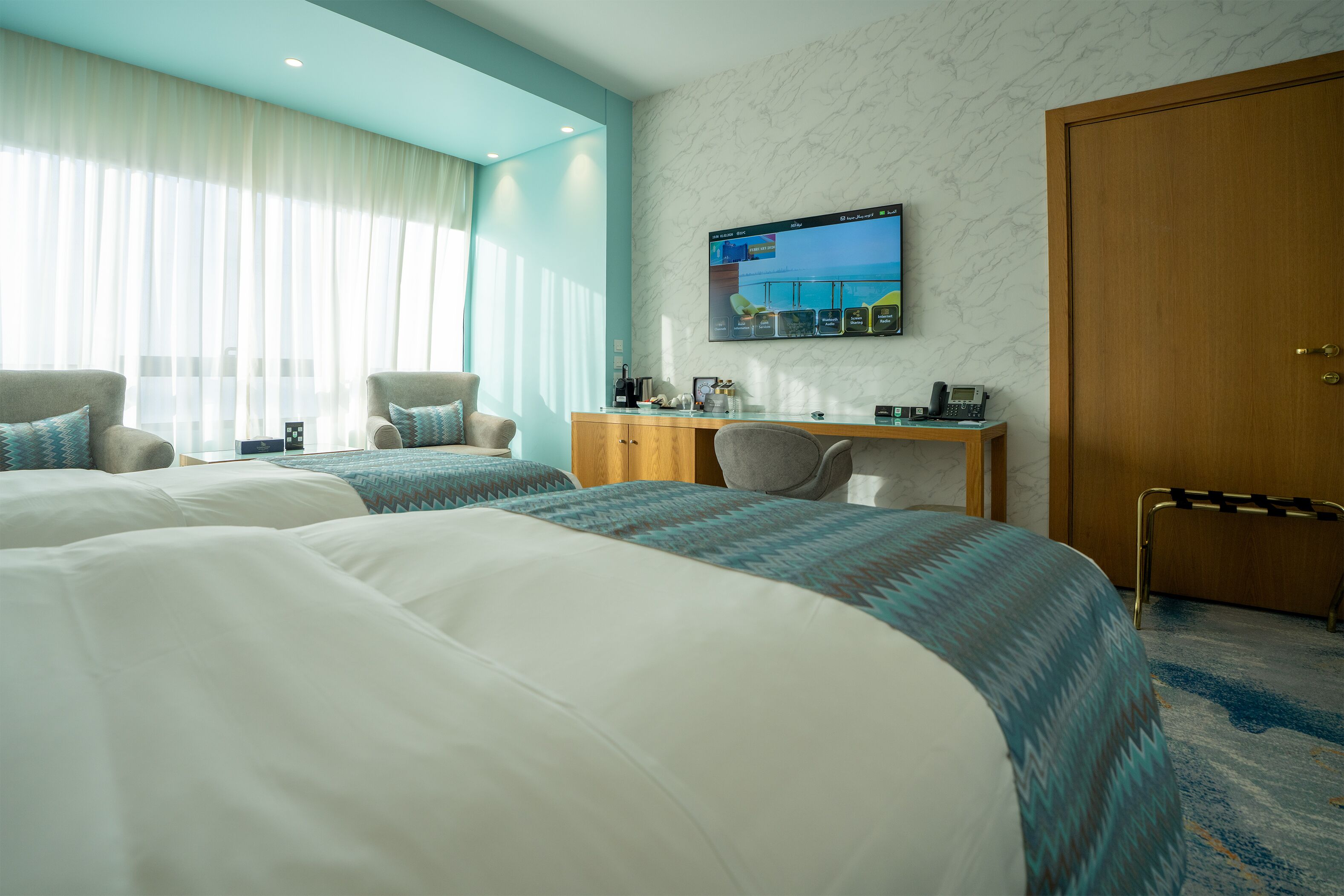 A hotel room with two beds, a desk, a chair, a TV, and a door.