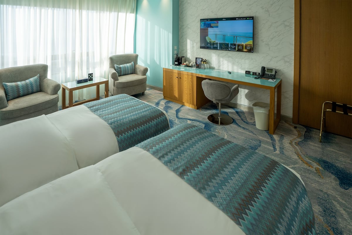 A hotel room with two beds, a desk, a chair, a TV, and a view of the sea.