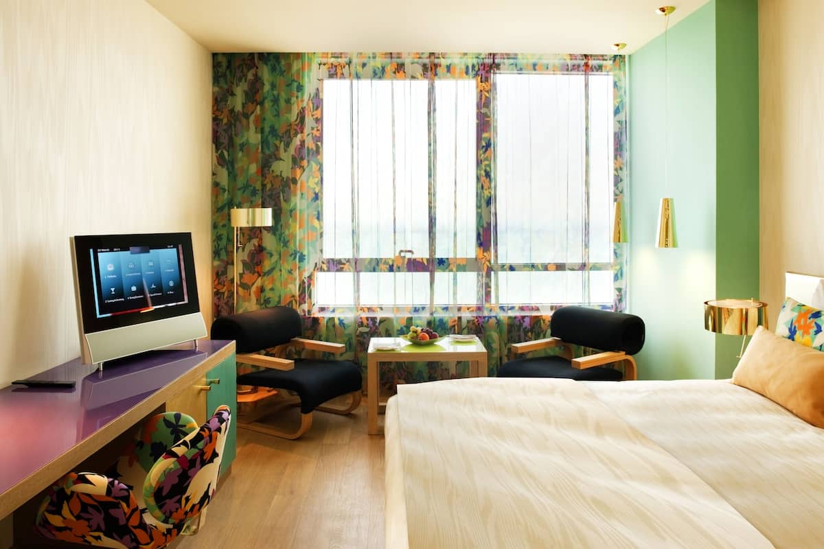 A hotel room with a bed, a desk with a computer, two chairs, a small table with a fruit bowl, and a window with curtains.