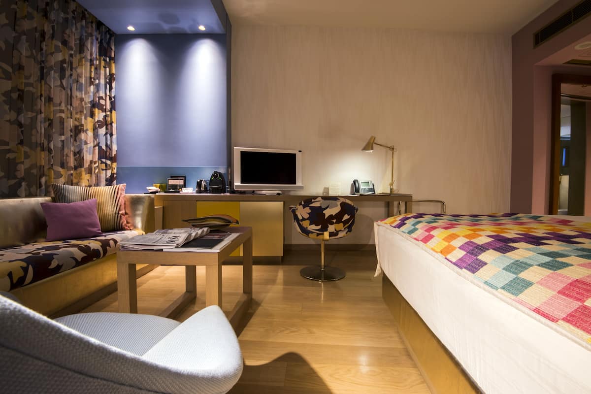 A modern hotel room with a sofa, a desk with a computer, a chair, and a bed with a colorful blanket.