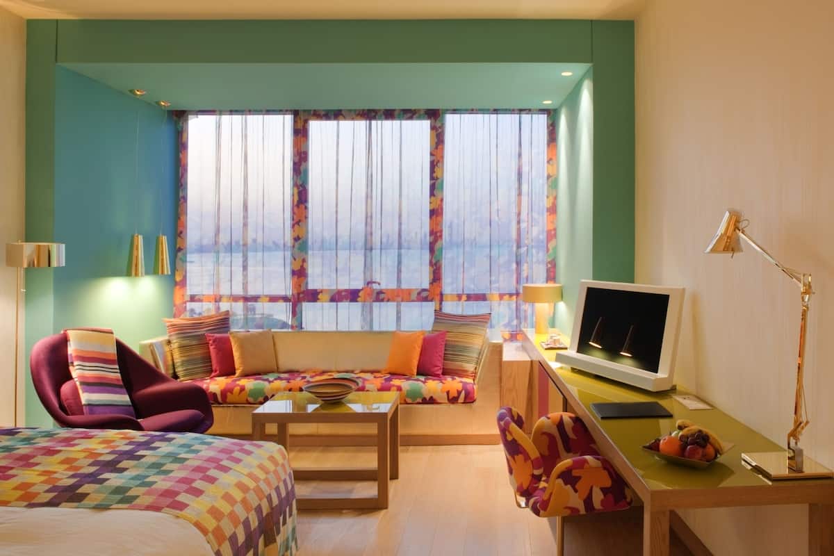 A hotel room with a large window, a sofa, a bed, a desk with a computer, and a chair.
