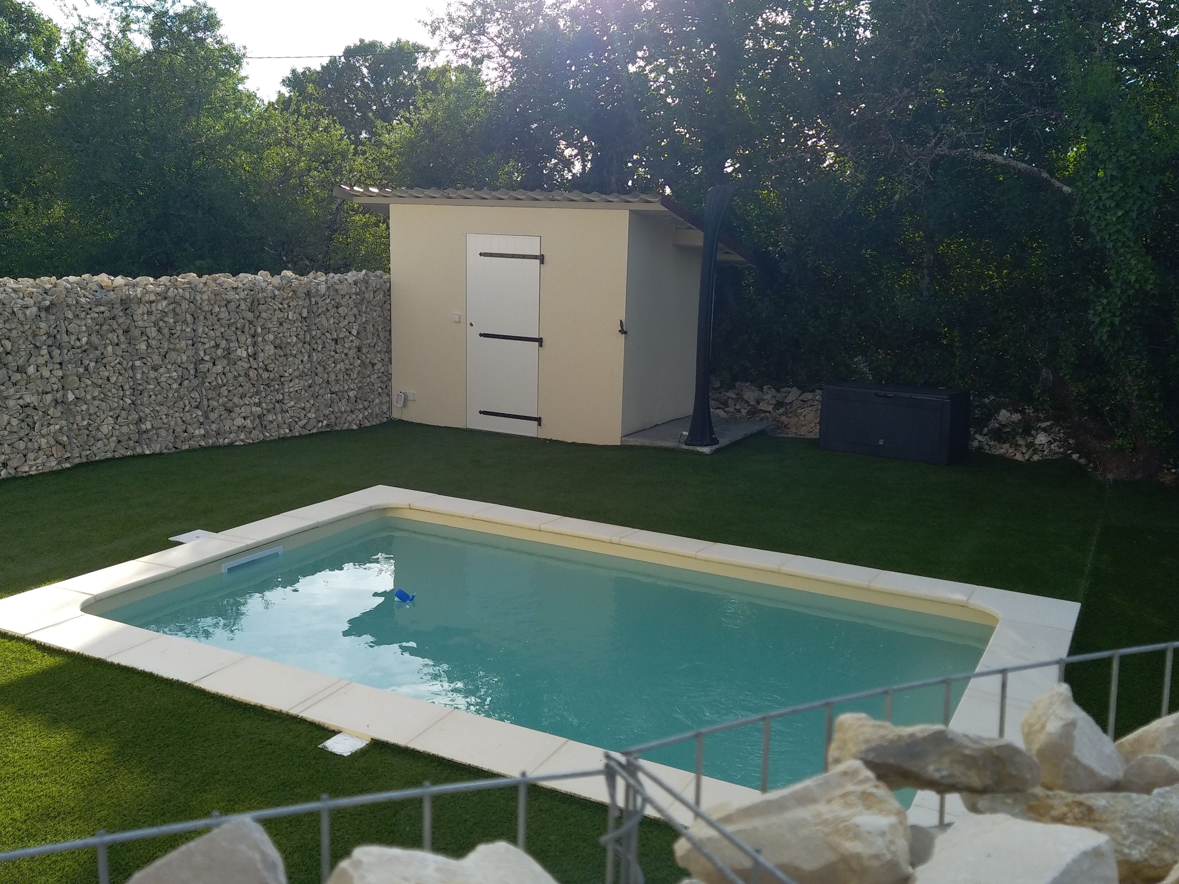 A rectangular swimming pool with a stone border, surrounded by a stone wall and a hedge.