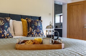 Un lit parfaitement fait, avec un plateau contenant divers éléments pour le petit-déjeuner, notamment des viennoiseries, du jus de fruits et du café, dans une chambre au design moderne, avec une cuisine ouverte visible.