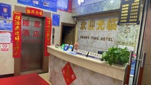 Un comptoir d’accueil d’hôtel avec une enseigne indiquant « Zhang Ying Hotel », un tapis rouge et une plante en pot.