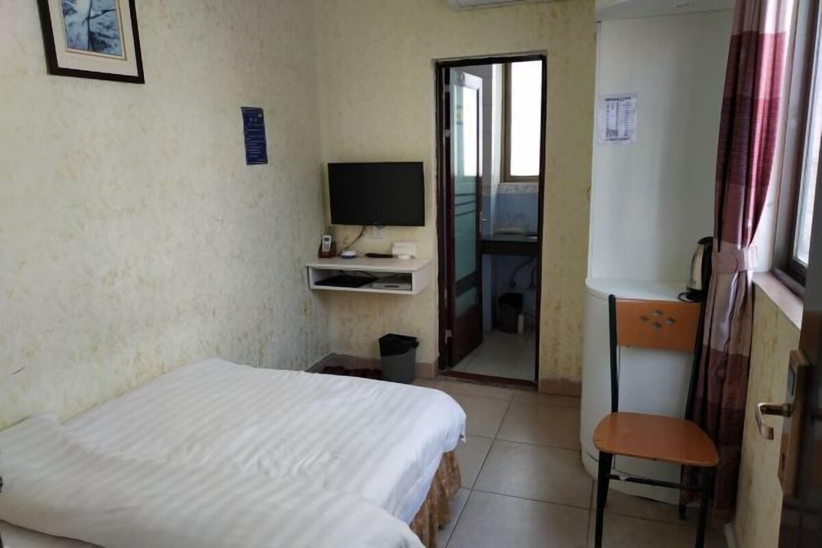A small, simple room with a bed, a desk with a computer, a chair, and a window with curtains.