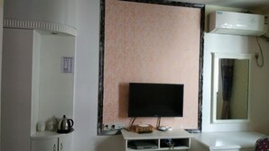 Une chambre dotée d’un téléviseur à écran plat fixé au mur, d’un meuble blanc contenant une bouilloire et des tasses, et d’une petite étagère avec une boîte et des télécommandes.