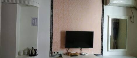 Una stanza con una televisione a schermo piatto montata a parete, un mobiletto bianco con bollitore e tazze, e un piccolo ripiano con una scatola e telecomandi.