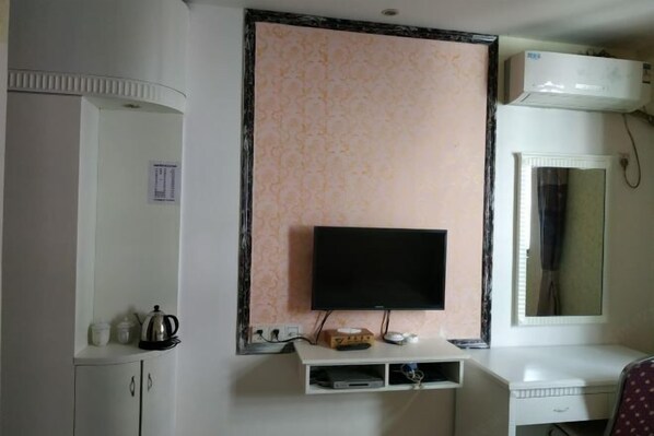 A room with a flat-screen TV mounted on a wall, a white cabinet with a kettle and cups, and a small shelf with a box and remote controls. - Zhang Ying Hotel (Dongguan)