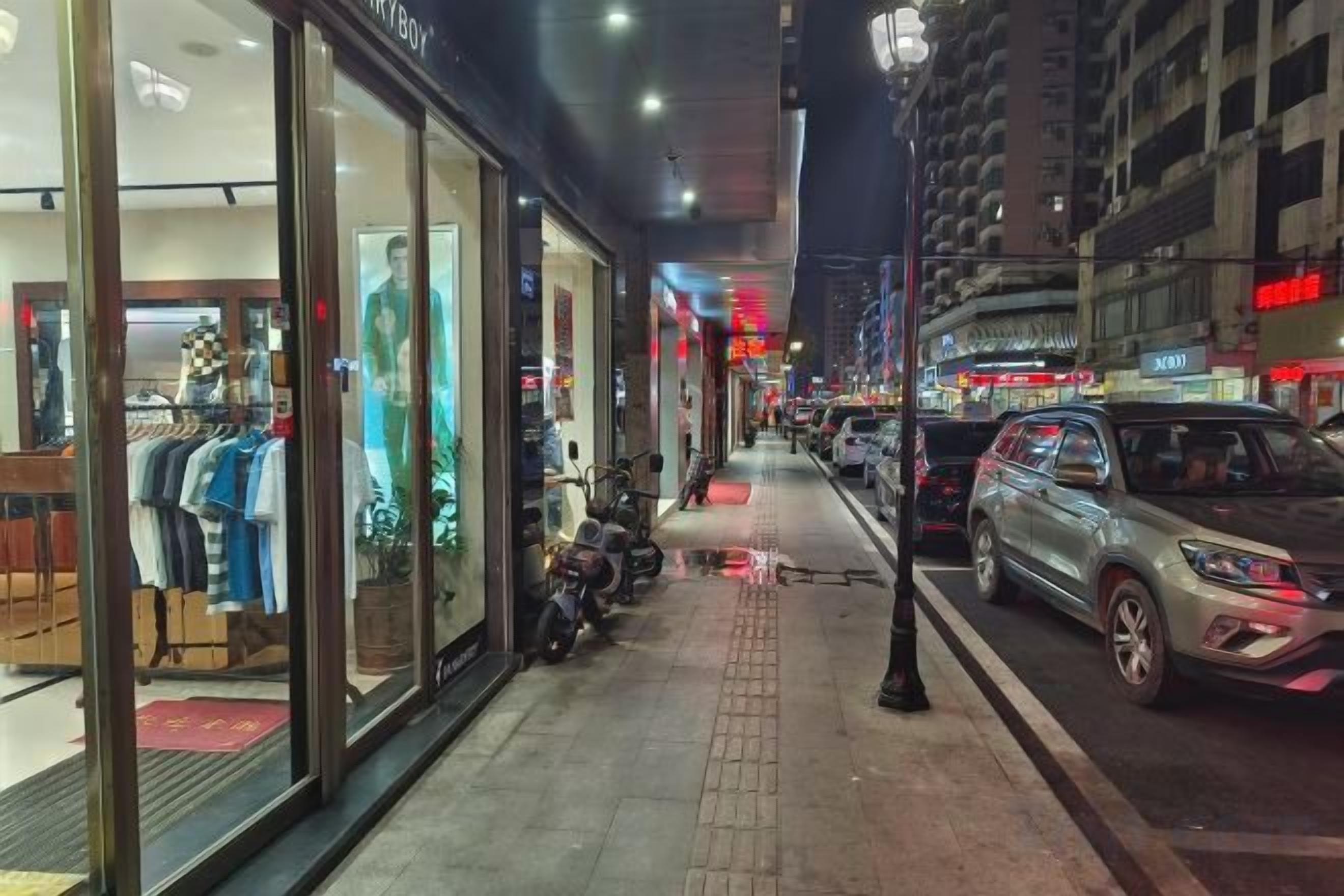 Vista nocturna de una calle con una tienda de ropa a la izquierda, motocicletas estacionadas y coches a la derecha.