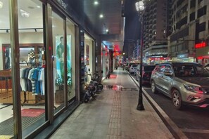 A nighttime street view with a clothing store on the left, parked motorcycles, and cars on the right. - Zhang Ying Hotel (Dongguan)