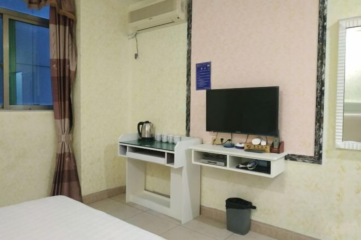 A hotel room with a bed, a TV mounted on the wall, a small desk with a kettle, cups, and a trash bin.