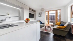 A modern kitchen with white cabinetry, a central island, and a fruit bowl. A living area with a sofa and coffee table is visible through an open door. - Luxury Penthouse Near Notting Hill Gate | Stunning Sunset Views (London)