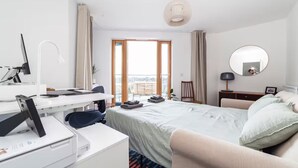 A modern hotel room with a large bed, a desk with a computer, a chair, a small table, a mirror, and a view of the city through the window. - Luxury Penthouse Near Notting Hill Gate | Stunning Sunset Views (London)